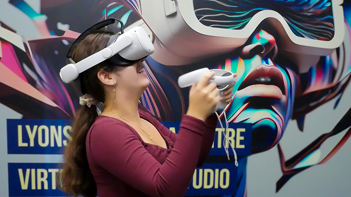 A young woman wears a virtual reality or VR headset and holds controllers in her hands while looking upward. Behind her, a large artistic mural covering an entire wall can be seen featuring an illustration of a futuristic woman wearing a VR headset.