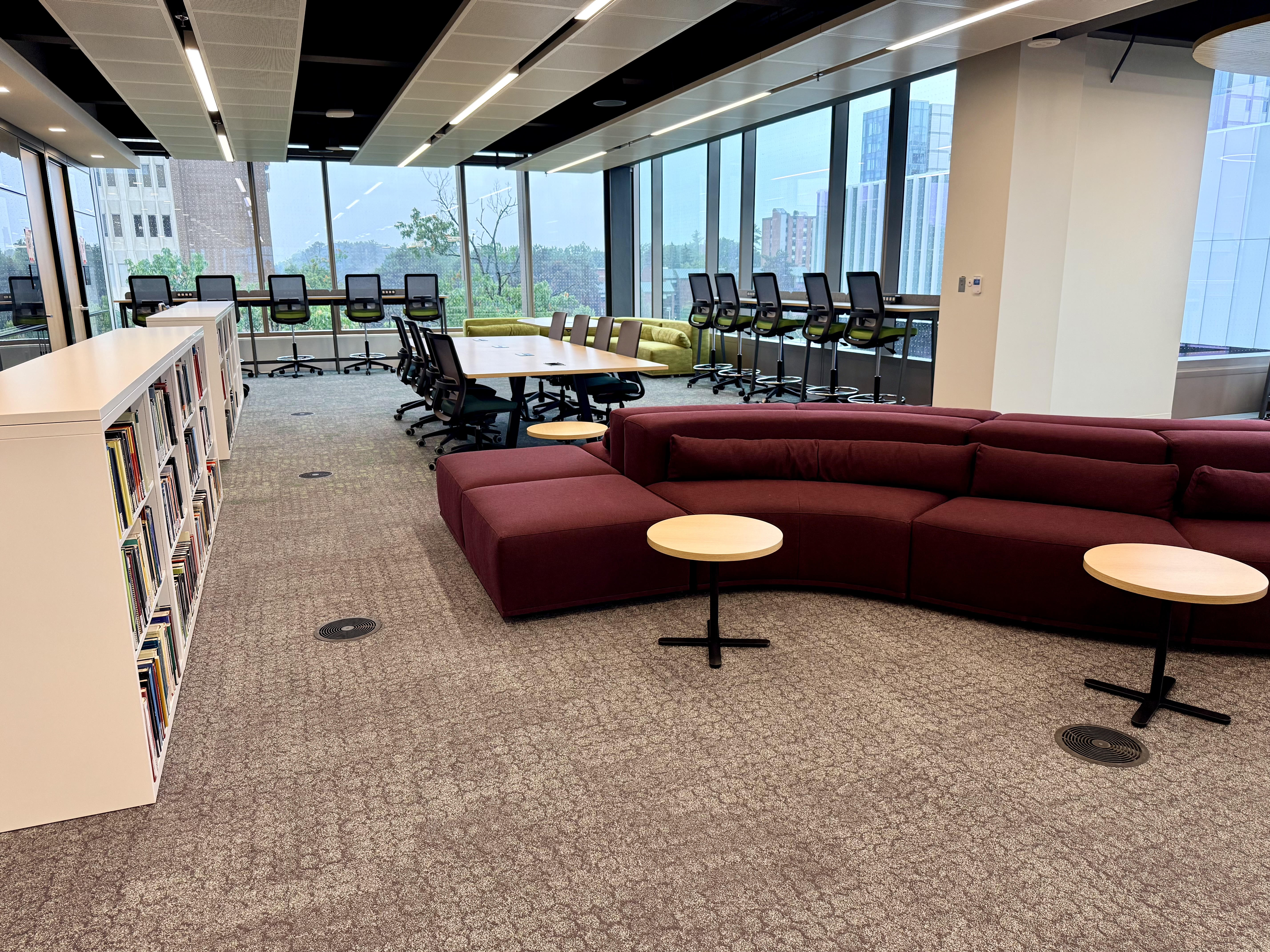 Innis Library Quiet Learning Zone with a bookshelf on the left and maroon plush seating with an an assortment of low lounge, standard, and high-top study tables by a window-lined wall..