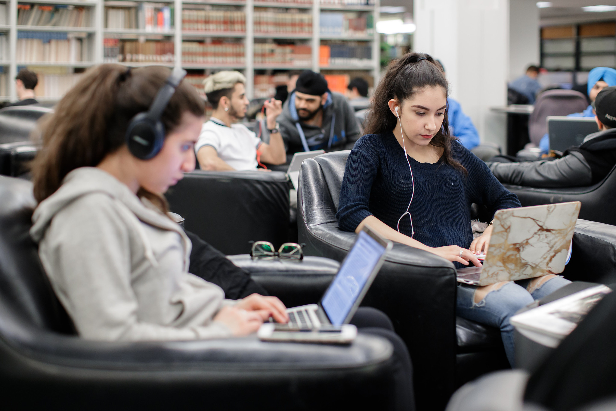 An image of two students sitting on chairs working on their laptops.